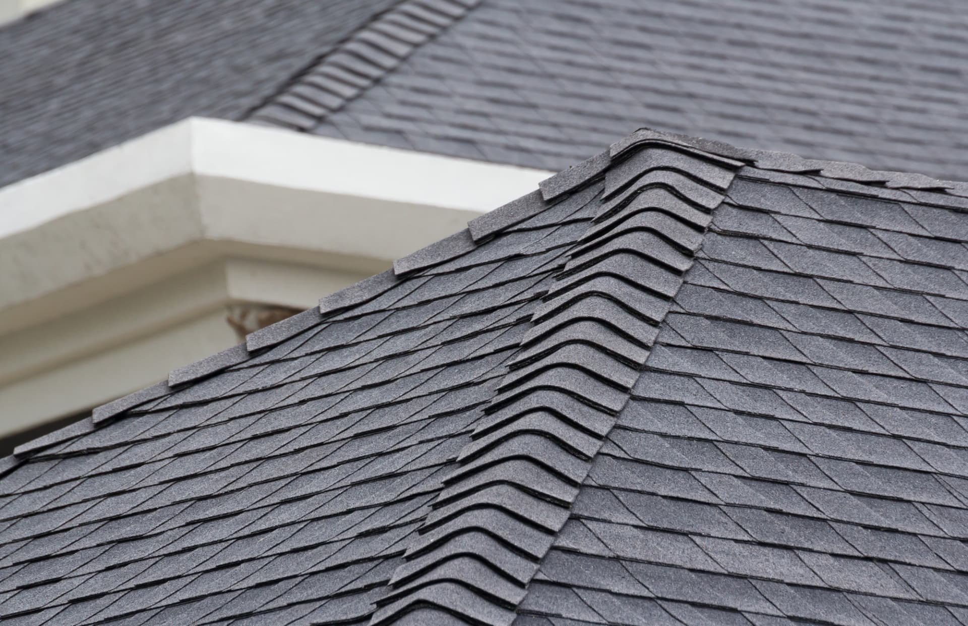 Close-up of grey asphalt shingles meeting at a hip, with ridge cap shingles running down the corner and white fascia behind.
