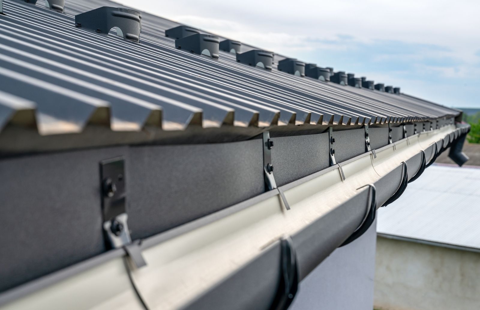 A pale gutter running along the eave of a dark corrugated metal roof, held in place by metal brackets.