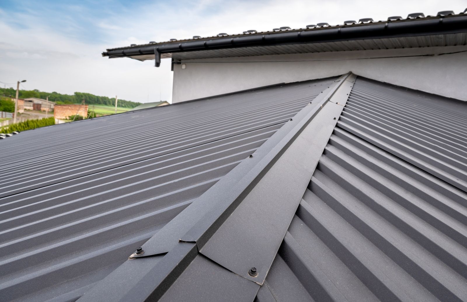 Dark grey ribbed metal roof panels meeting at a capped ridge, fixed with exposed fasteners, with a gutter along the eave above.